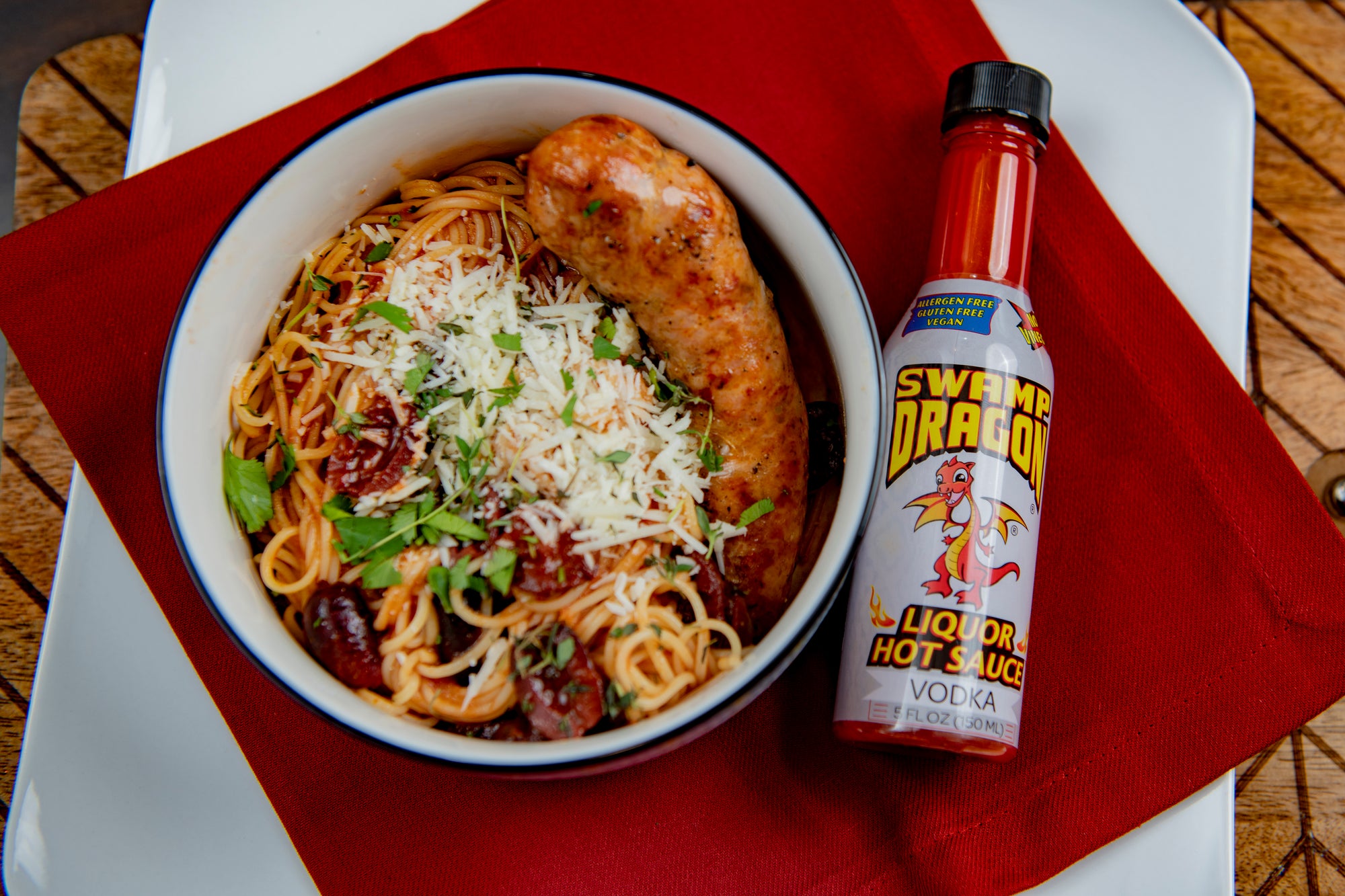 Bowl of spaghetti with red sauce, a link of italian sausage, and shredded parmesan cheese. A 5 ounce bottle of Swamp Dragon "Vodka Dragon Hot Sauce" sits next to the bowl.