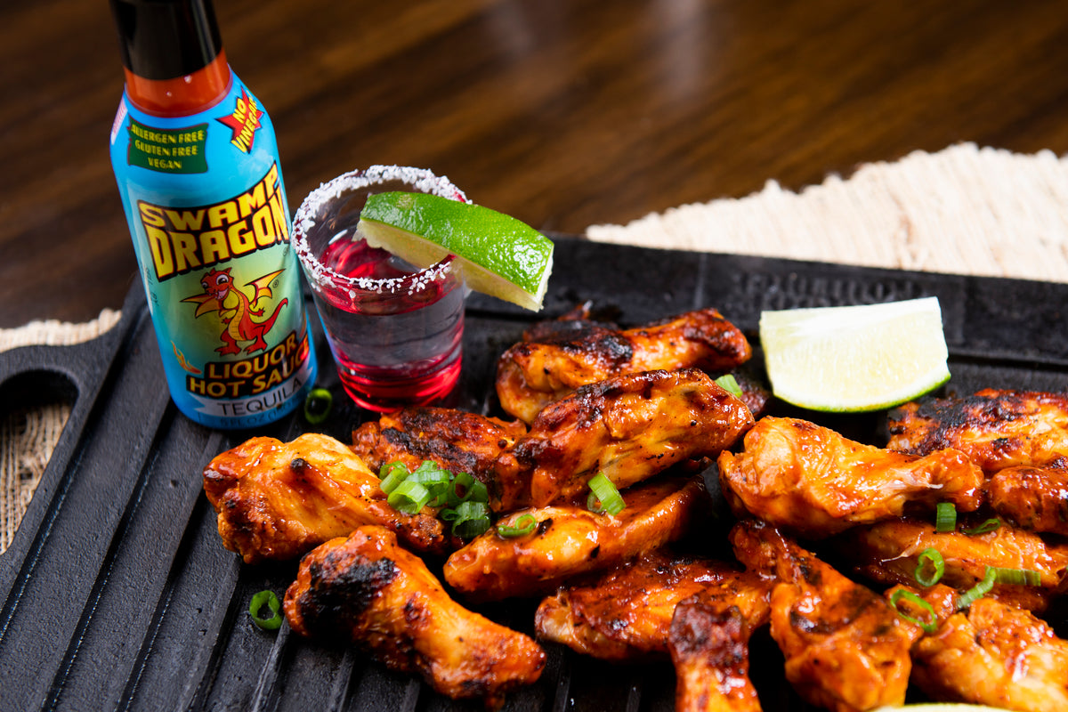A bottle of Swamp Dragon Tequila Hot Sauce next to a pile of tequila chicken wings and a shot of tequila with salt and lime wedge