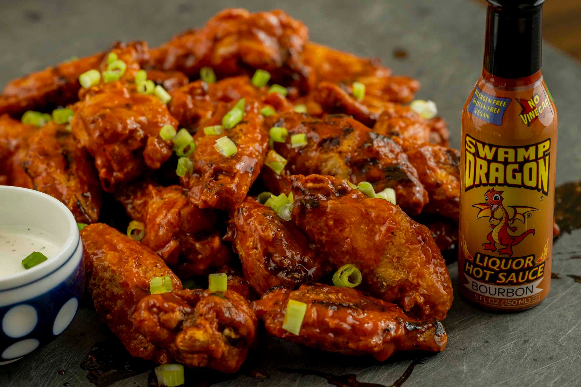 A pile of grilled chicken wings covered in hot sauce, garnished with green onions, with a bottle of Swamp Dragon Bourbon Hot Sauce