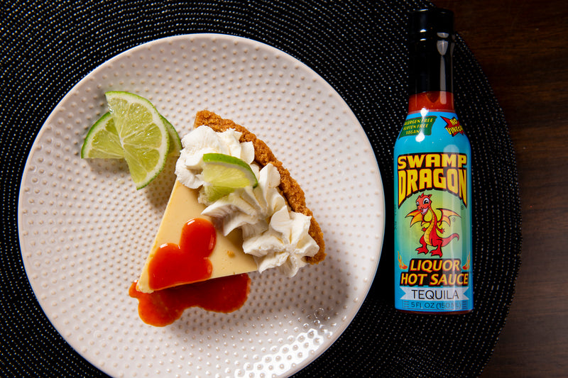 A slice of key lime pie with lime wedge garnish, whipped cream, and a splash of Swamp Dragon Tequila Hot Sauce next to it. 