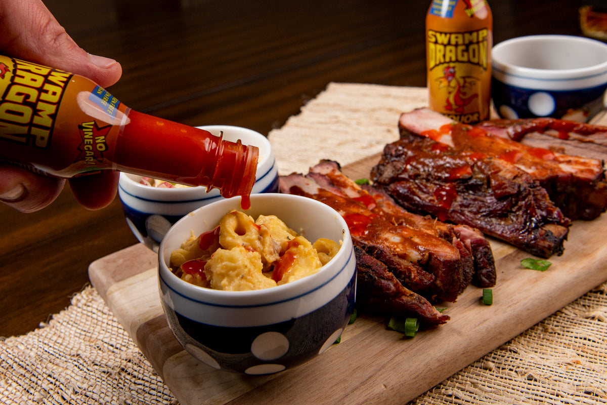 Swamp Dragon Bourbon Hot Sauce pouring out onto macaroni and cheese with a plate of barbecue ribs