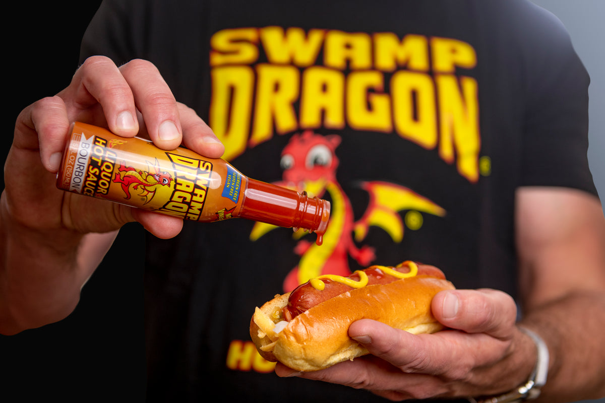 Swamp Dragon Bourbon Hot Sauce pouring onto a hot dog with Swamp Dragon T-shirt in background