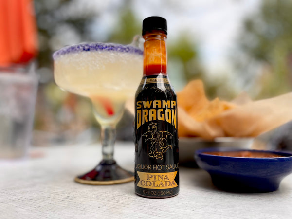 A five ounce bottle of Swamp Dragon Pina Colada Hot Sauce sitting in front of a margarita