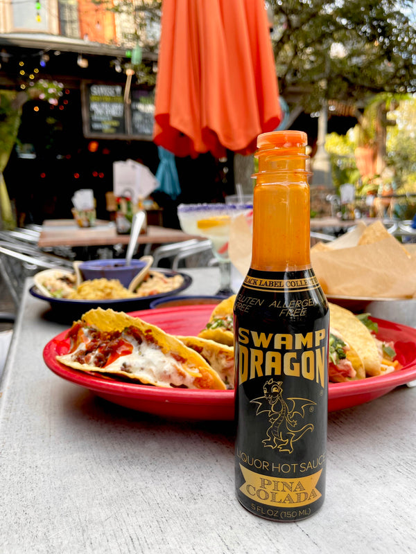 A five ounce bottle of Swamp Dragon Pina Colada Hot Sauce on a table in front of a plate of tacos